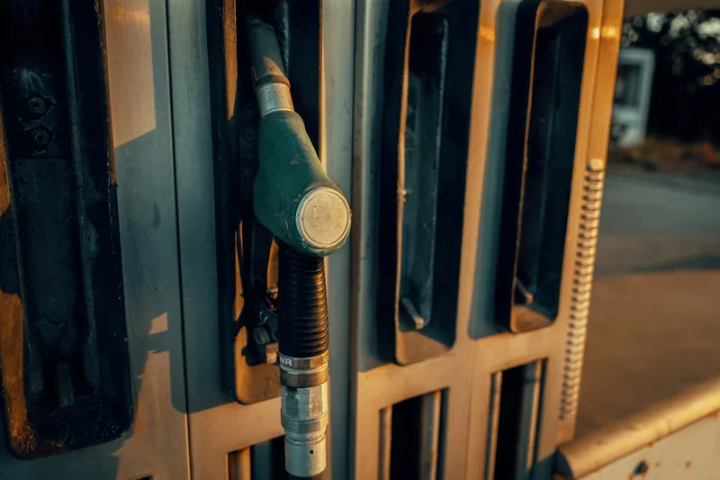 Close-up of a green fuel pump nozzle resting in its holder on a forecourt pump, lit by low evening sun.